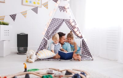 A father reads a book to his two young children inside a polka-dot play tent in a bright, cozy room, with toys and a stuffed animal nearby.