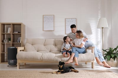 A happy family of three sits together on a beige sofa in a bright living room, smiling and holding a book, while a small black and brown dog lies on a rug in the foreground.
